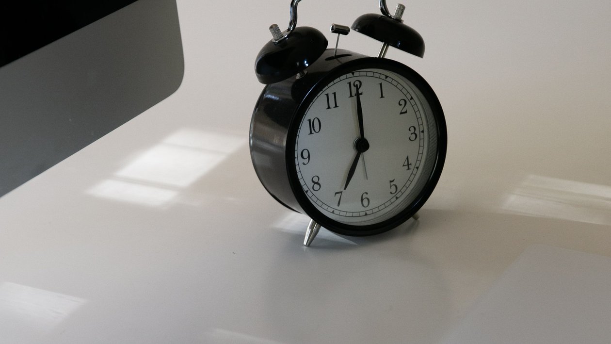 A black alarm clock sits on a white office desk beside the edge of a computer monitor.