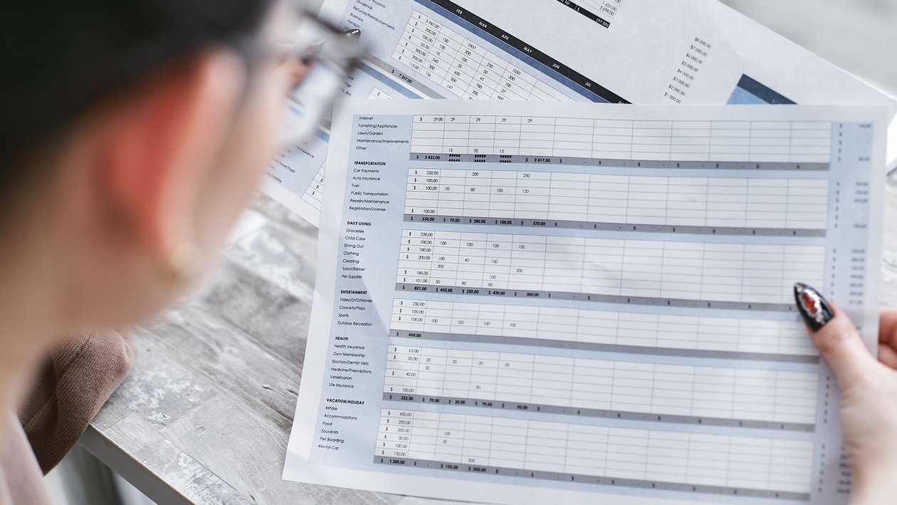 A person holds a printed spreadsheet full of figures and checks the rows against a report on the desk.