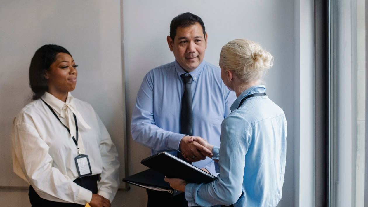 A manager shaking hands with a colleague holding folders while another colleague looks on in a bright office.