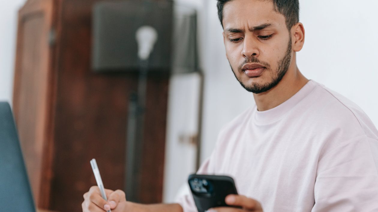 A young man at a desk frowning at his phone while holding a pen over a notebook, waiting on news.
