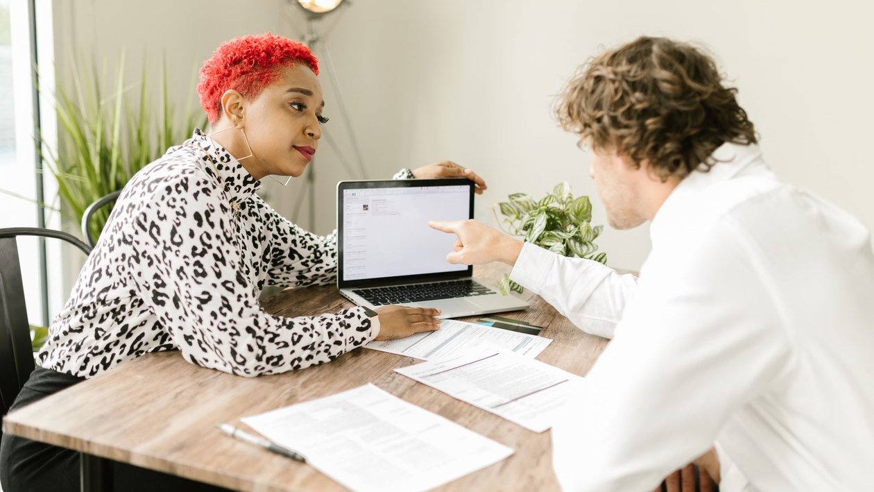 A candidate showing work on a laptop to an interviewer across a wooden table covered with printed documents.