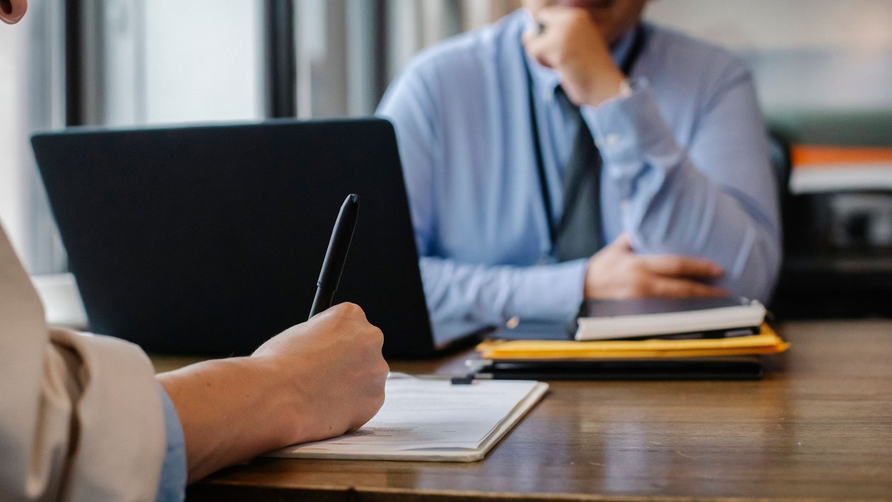 A job candidate writes answers on a notepad at a wooden desk while a manager behind a laptop watches.