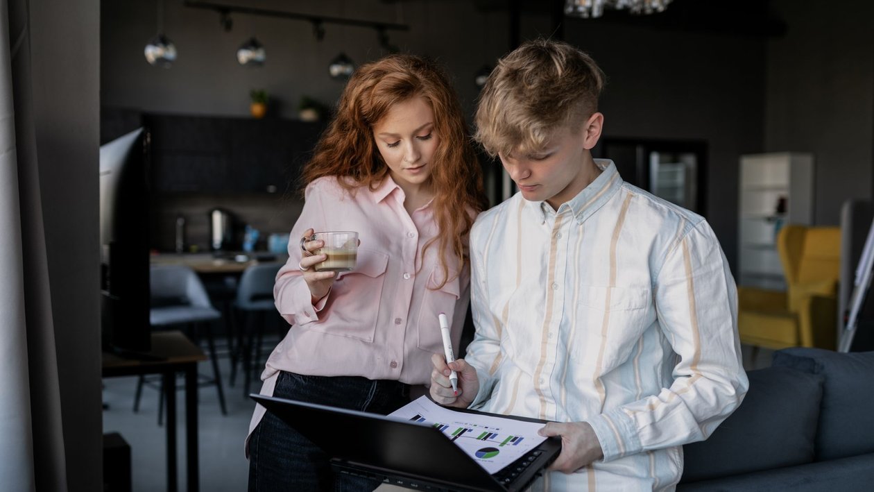A young man holds a laptop with chart printouts while a colleague holding a coffee looks on in a modern office.