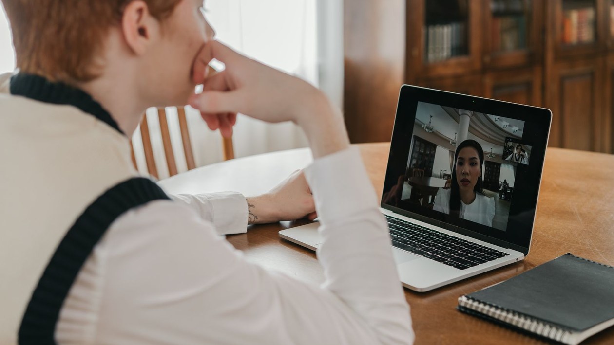 A person sits at a wooden table at home with a hand on their chin, watching a woman speak on a laptop video call.