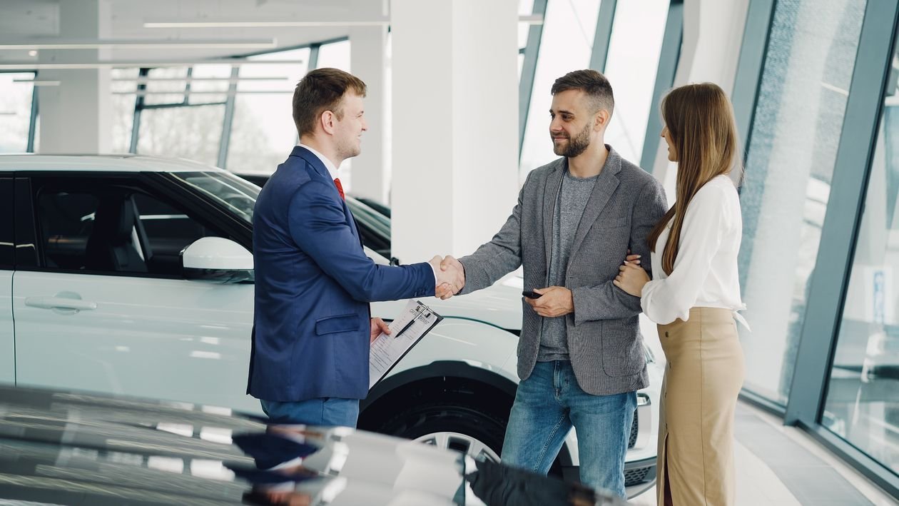 A car salesman holding paperwork shakes hands with a couple in a dealership showroom after a deal.