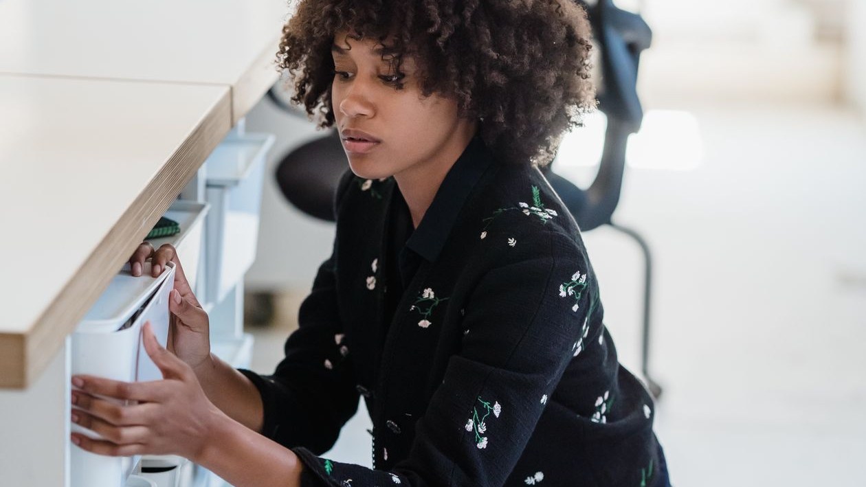 A woman in an office kneeling beside a desk and searching through files in a set of drawers.