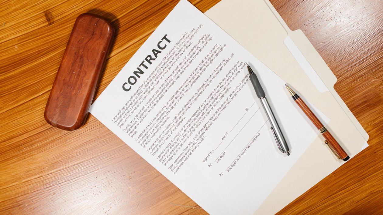 A printed contract with two pens and a pen case on a wooden desk.