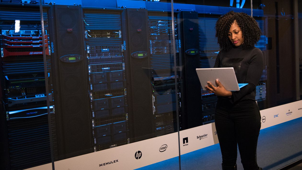 An engineer holding a laptop while monitoring rows of servers behind glass in a data center.