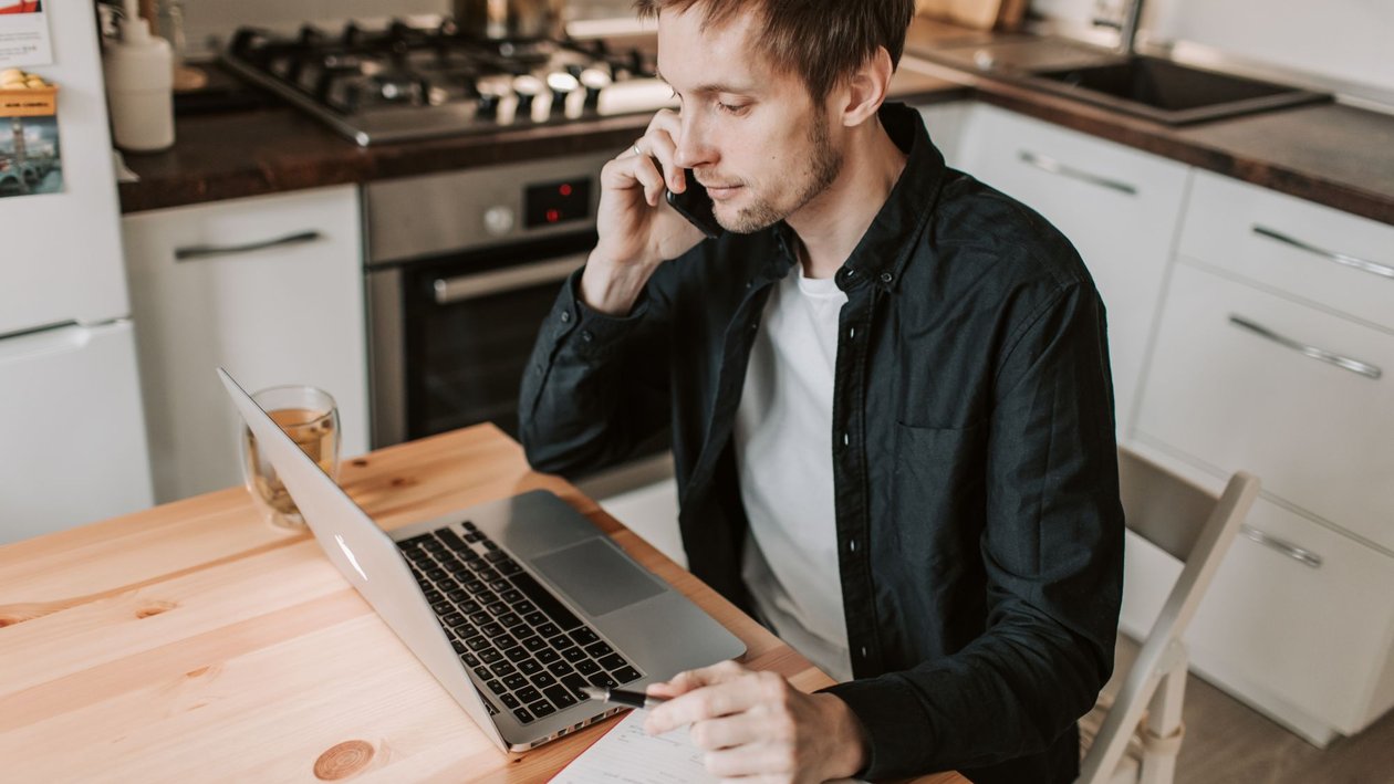 A young man talks on a phone while working on a laptop at a wooden kitchen table.