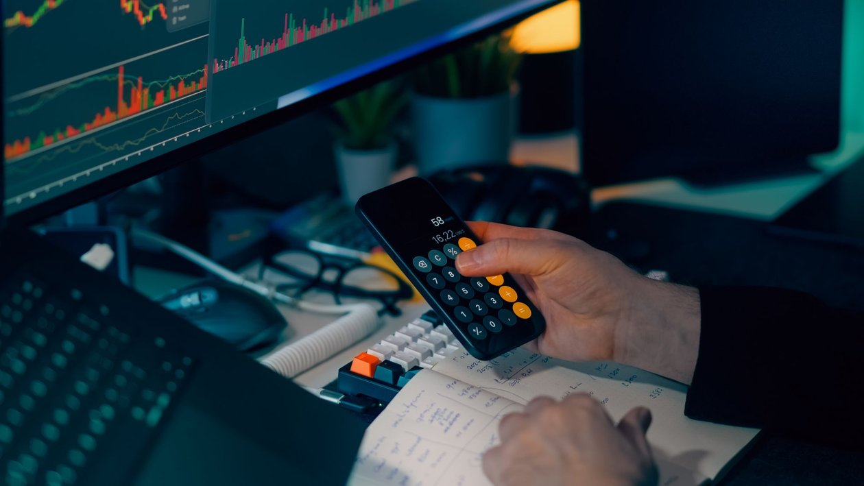 A person checking figures on a calculator and in a notebook beside a monitor showing financial charts.