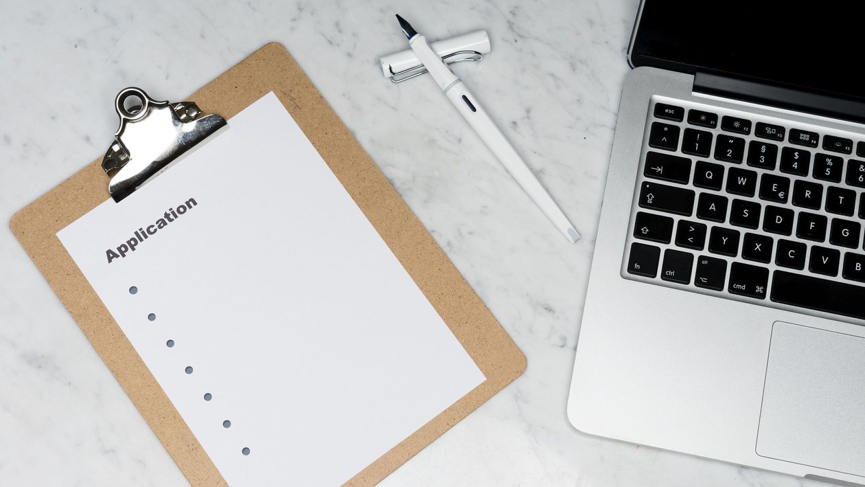 A clipboard holding a blank application form beside a pen and an open laptop on a marble desk.