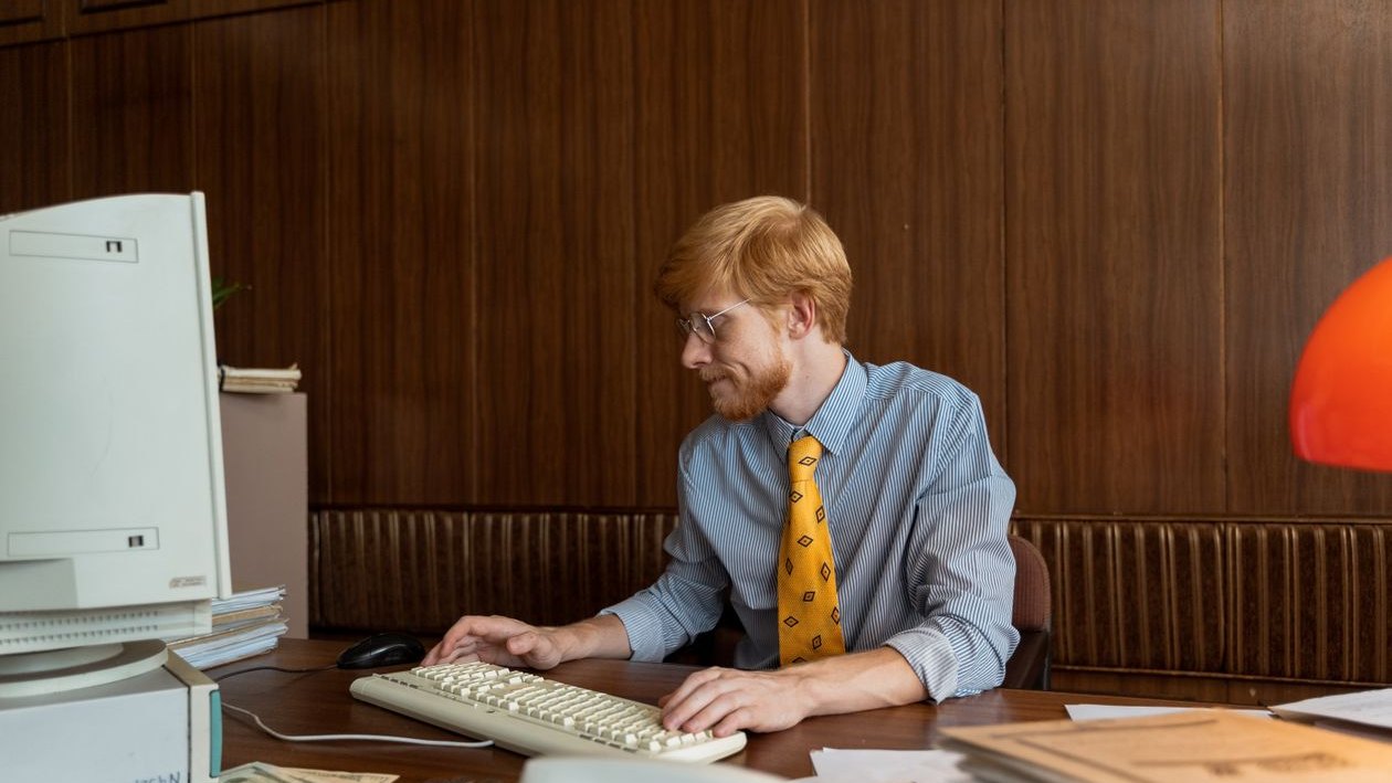 A man in a shirt and tie typing on an old beige desktop computer at a wood panelled office desk.