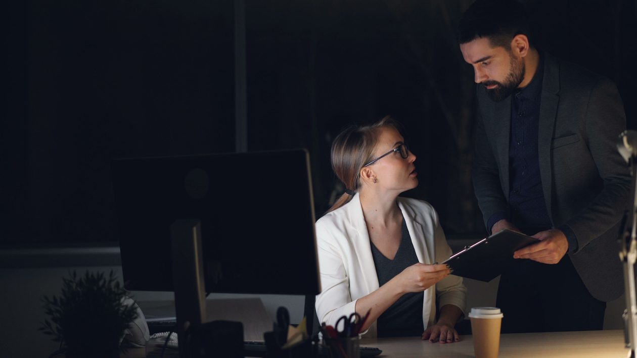 A manager hands a clipboard to an analyst at her desk in a dark office late at night.