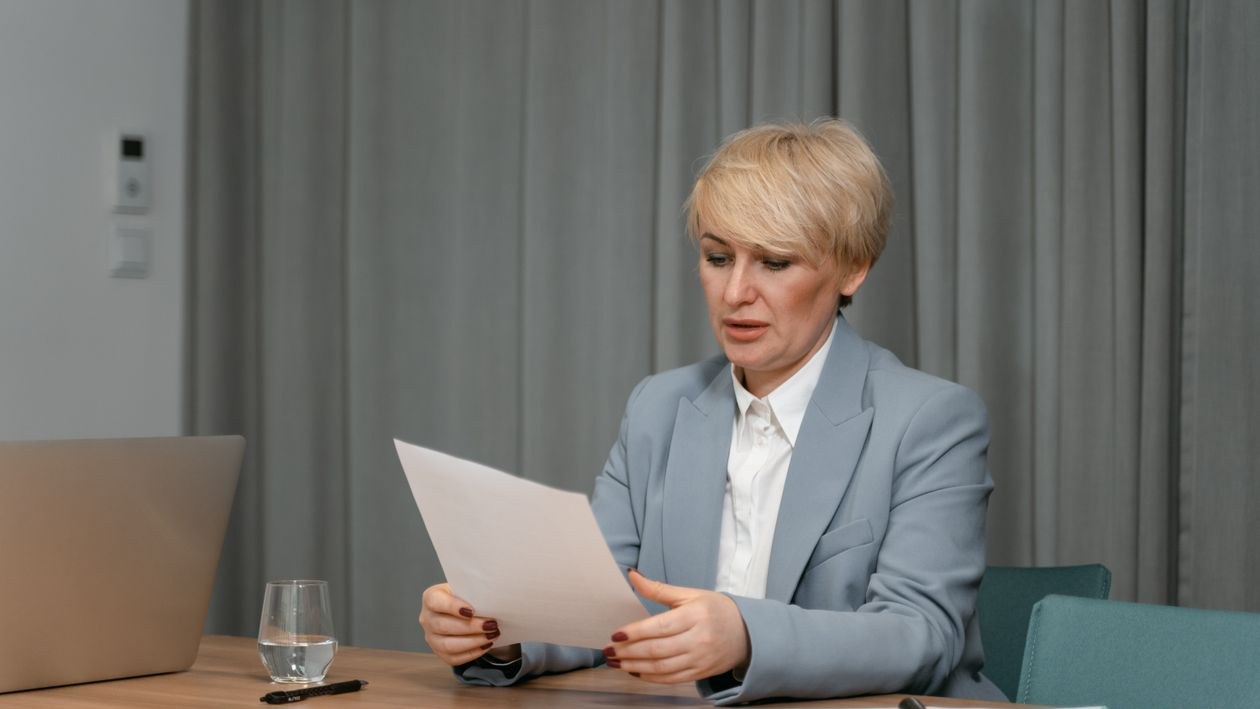 A manager in a light blue blazer reads a printed page at a meeting table next to a laptop and a glass of water.