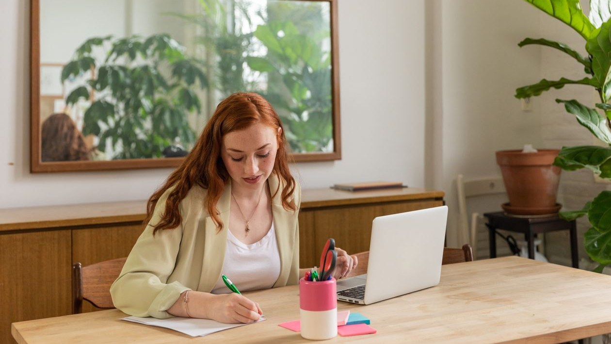A hiring manager writing notes at a wooden desk beside an open laptop in a bright office with plants.
