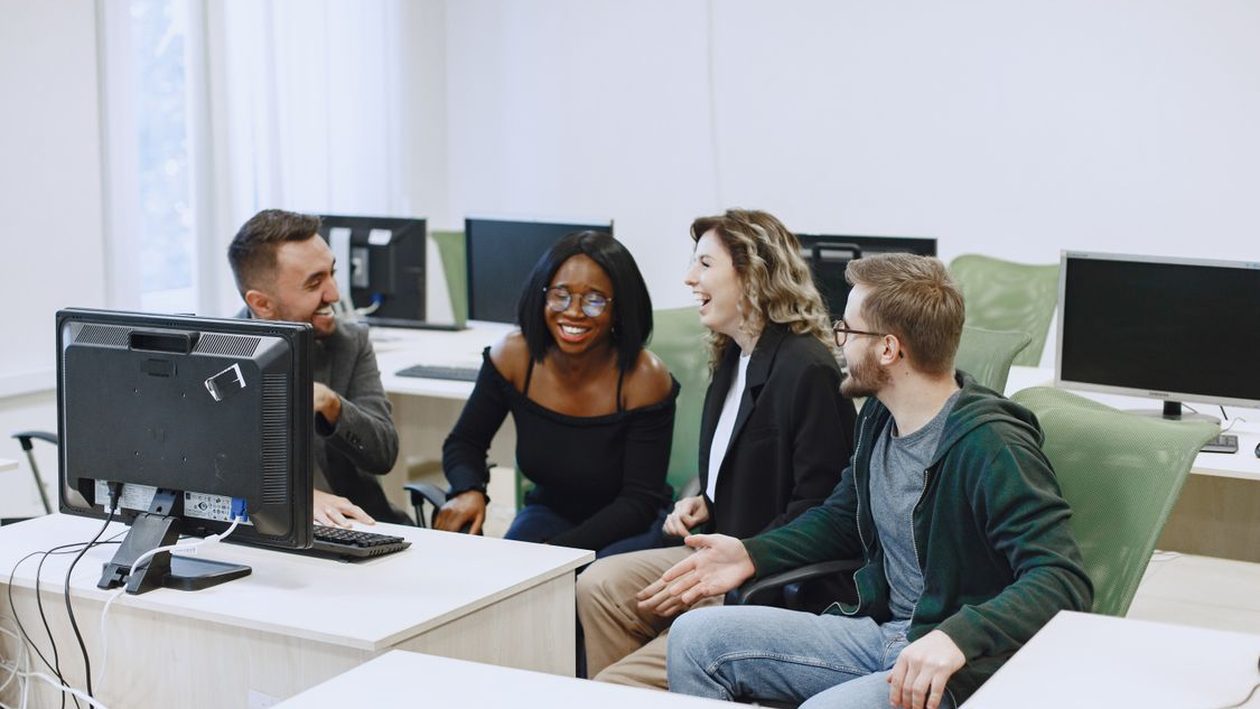 Four colleagues laughing and working together around a desktop computer in a bright office.
