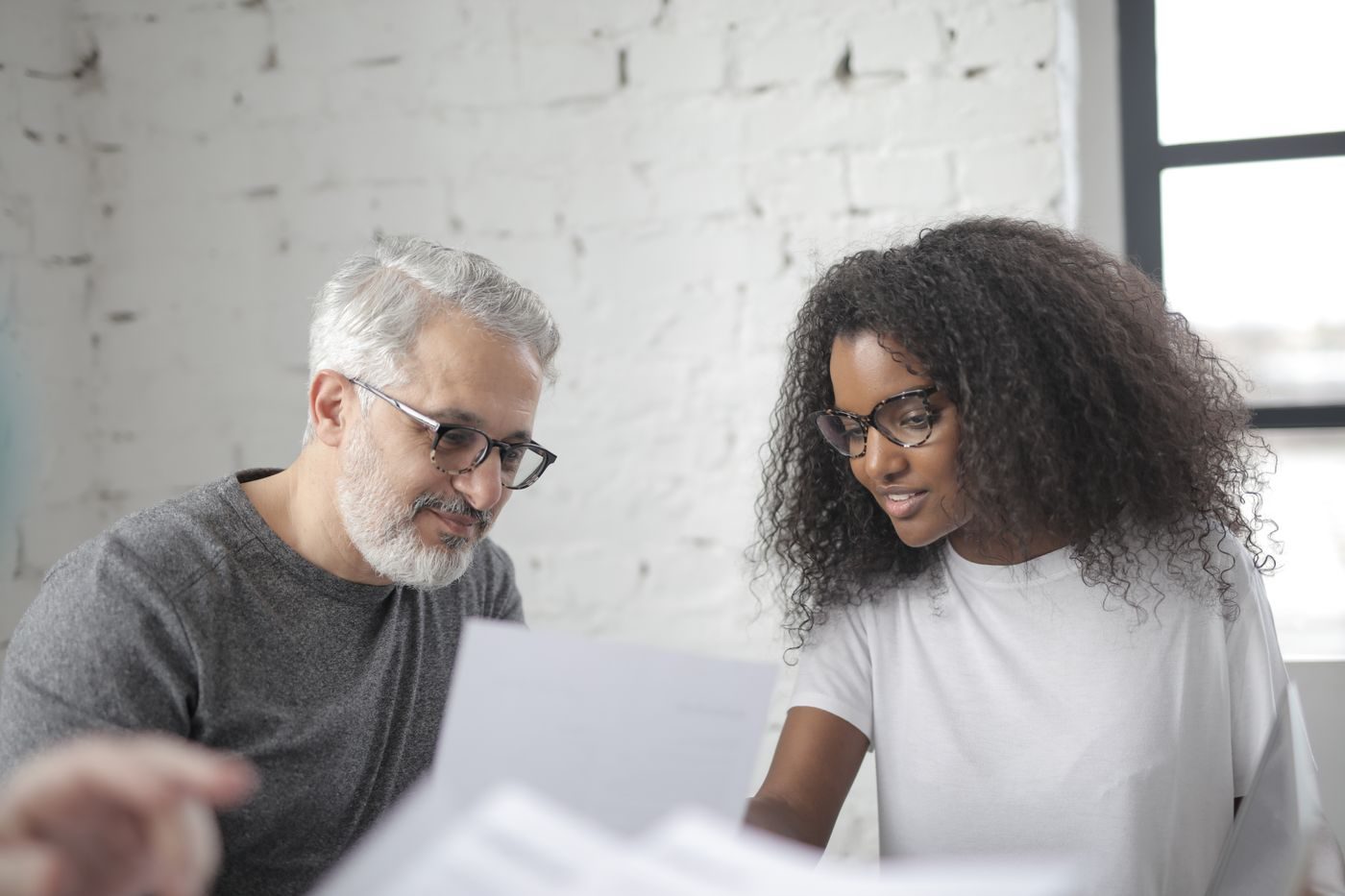 A new employee in glasses reads printed papers beside a senior colleague in a bright office.