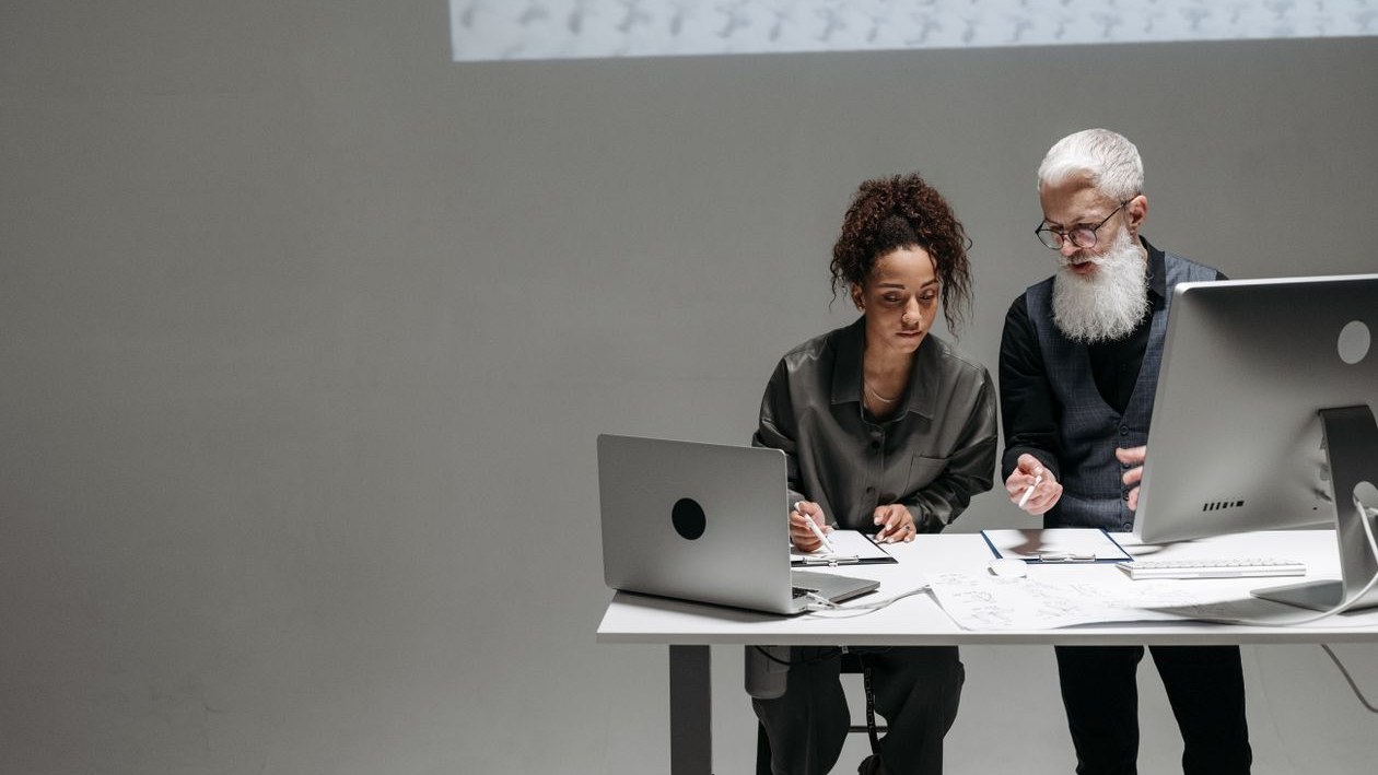 An older man with a white beard points at papers while a younger colleague takes notes beside a laptop and a desktop monitor.