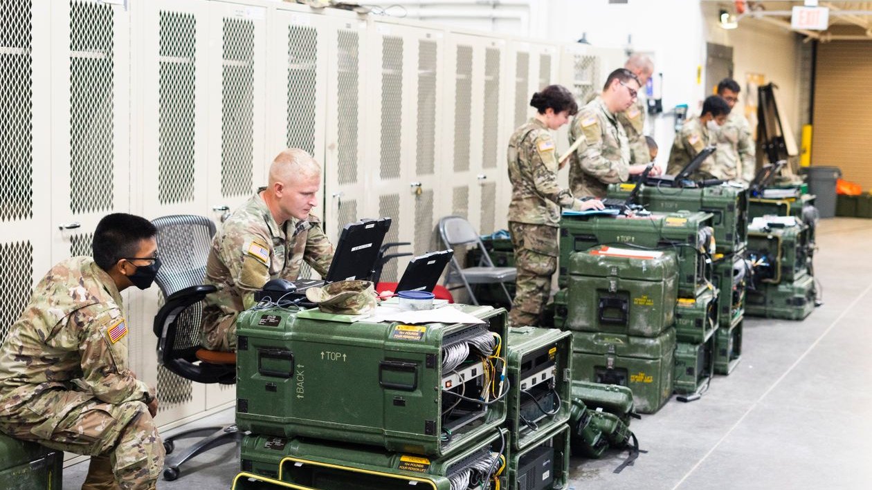 Soldiers in uniform working on laptops set on equipment cases in a long indoor workspace.