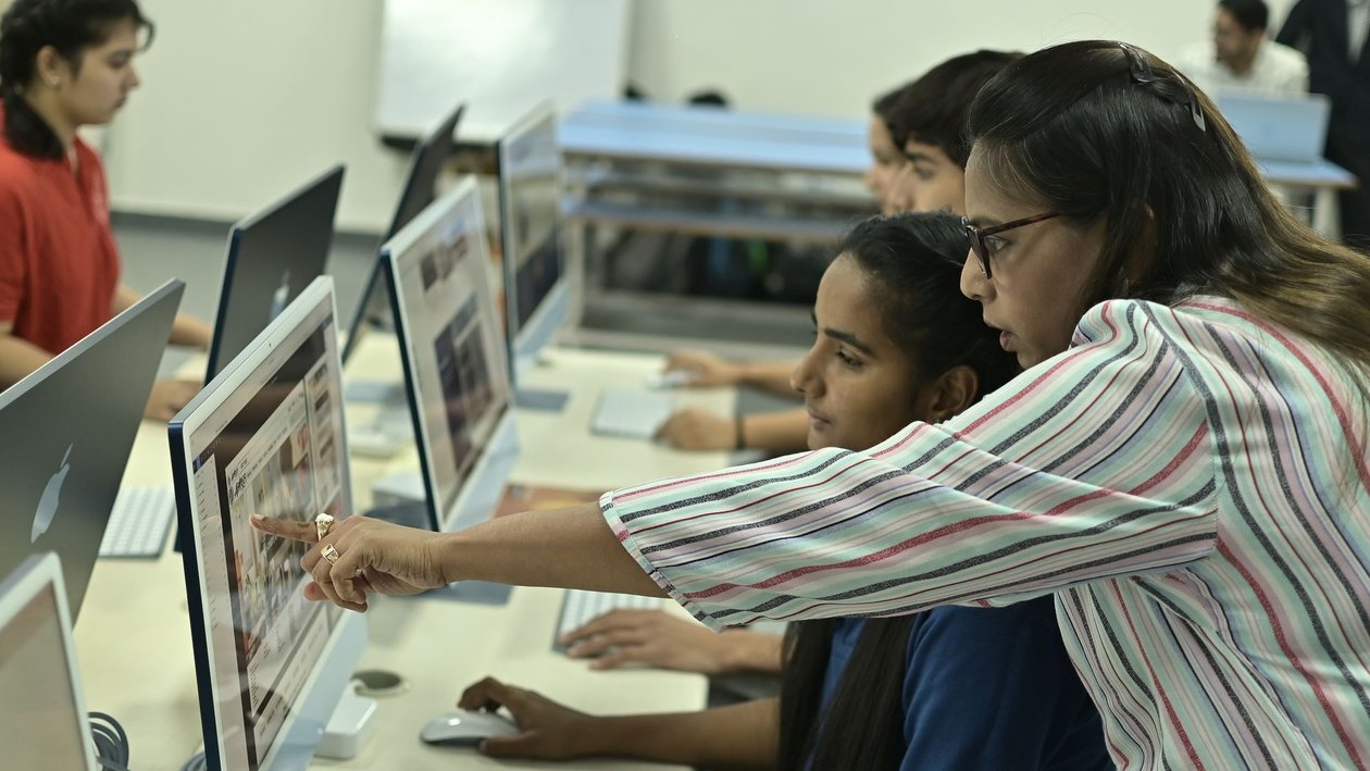 A teacher leans in and points at a computer screen while students work at desktop computers in a computer lab.