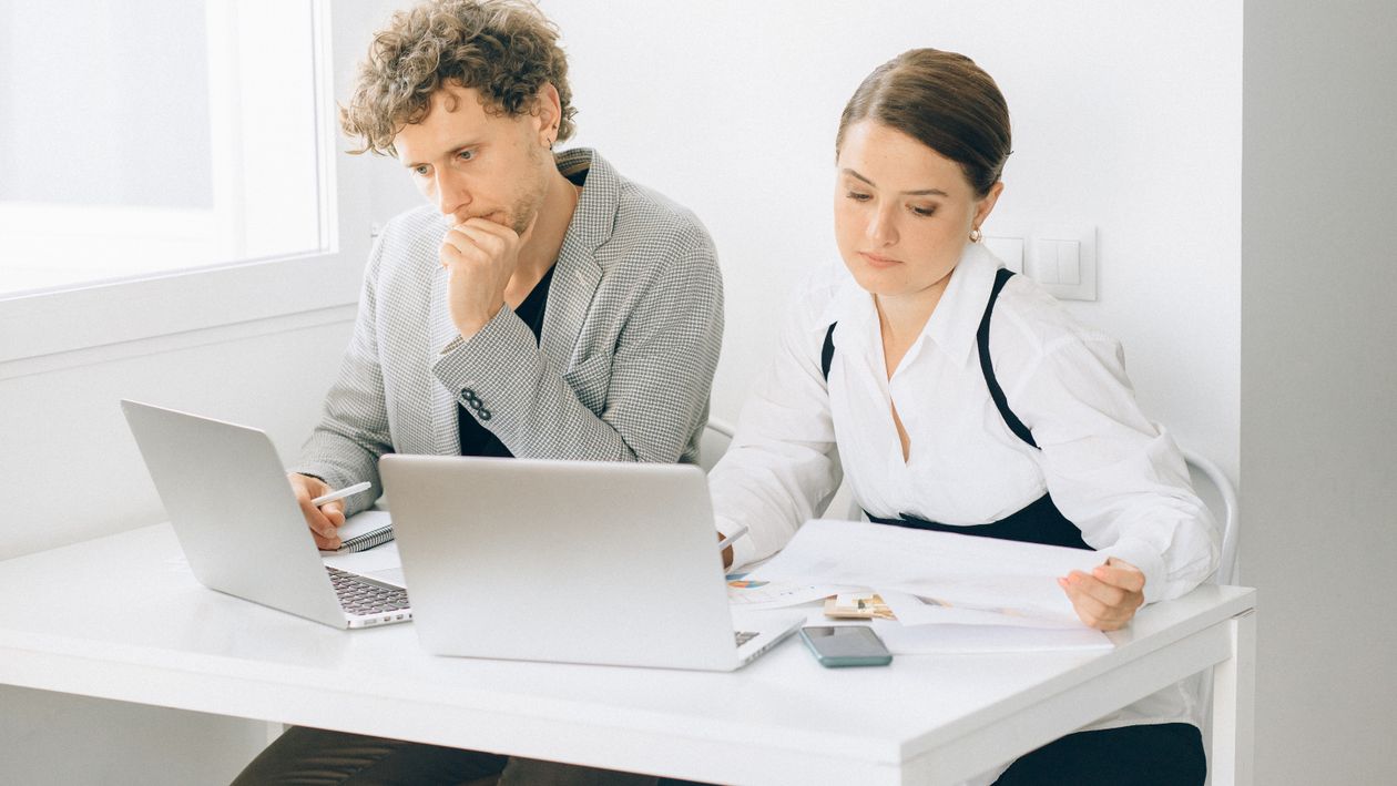 Two coworkers at a white desk with two laptops, one reading printed documents, reviewing paperwork together in a bright office.