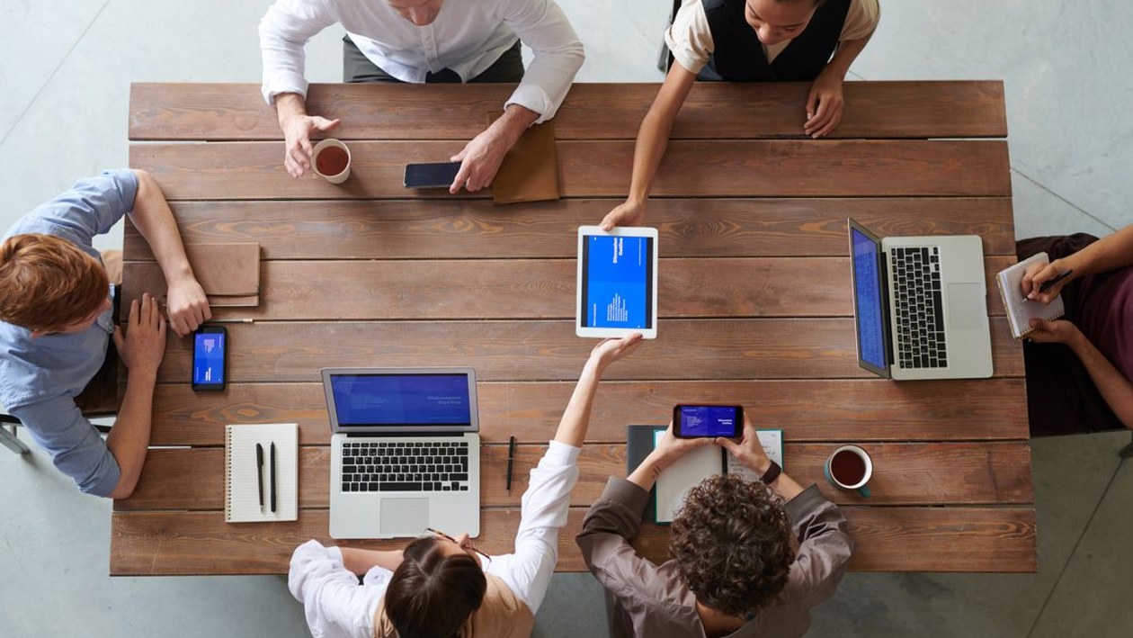 A team seated around a table reviewing work together on laptops, a tablet, and phones, seen from above.