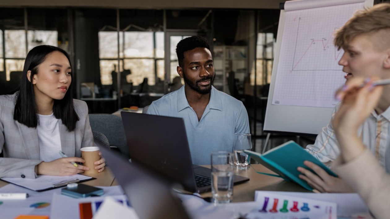 A team discussing work around a table with laptops, charts, and notes in a modern office.