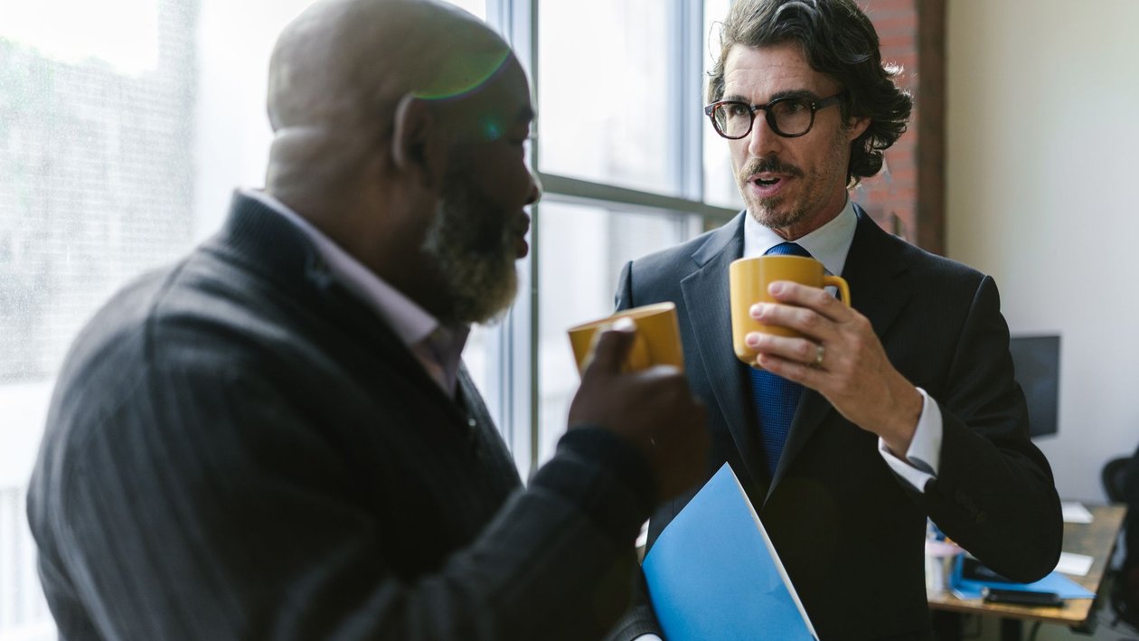 Two men in business clothes stand by an office window holding coffee mugs and talking.