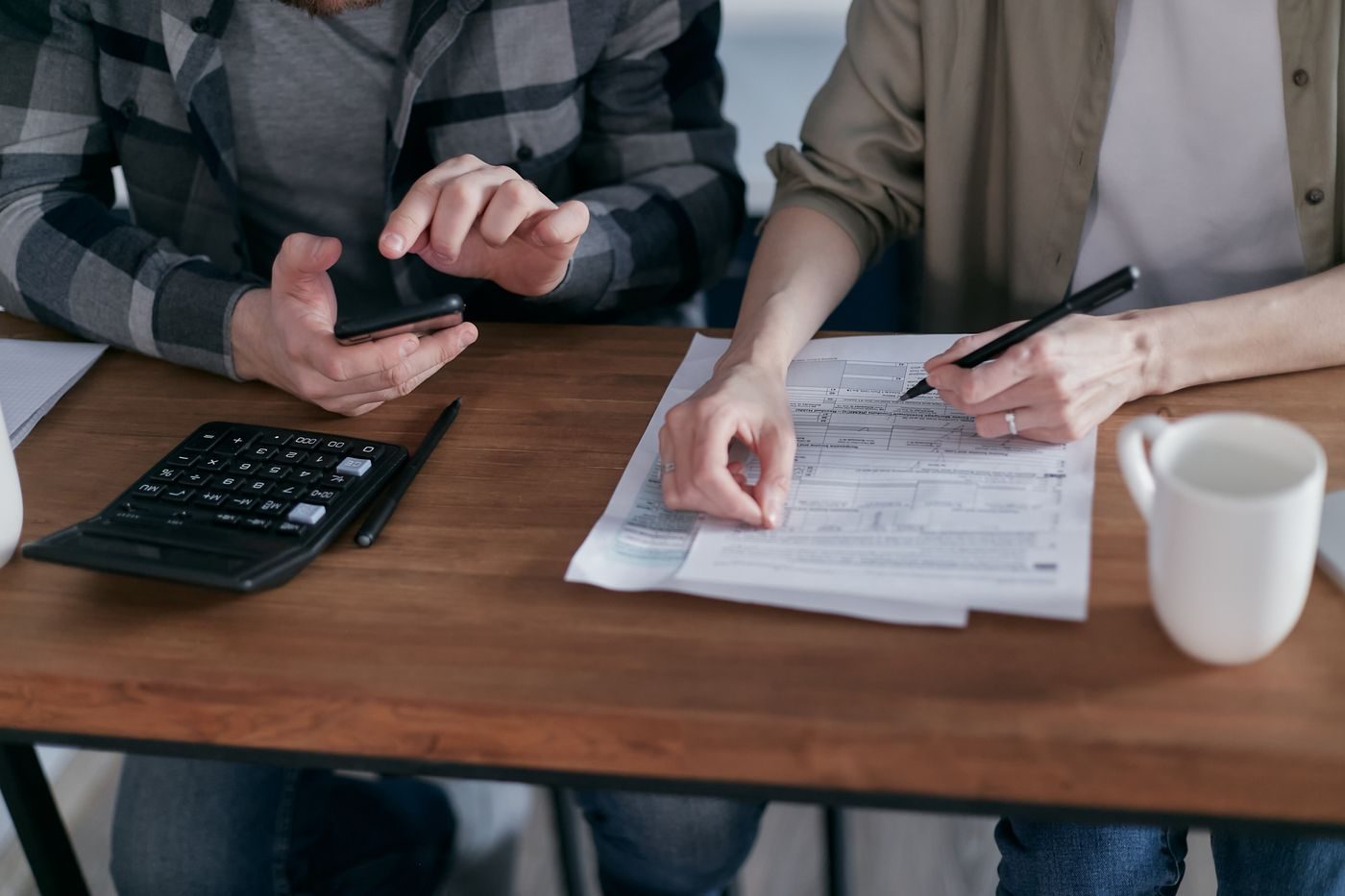 Two people at a wooden table go over tax paperwork with a pen, a phone, a calculator and a coffee mug.