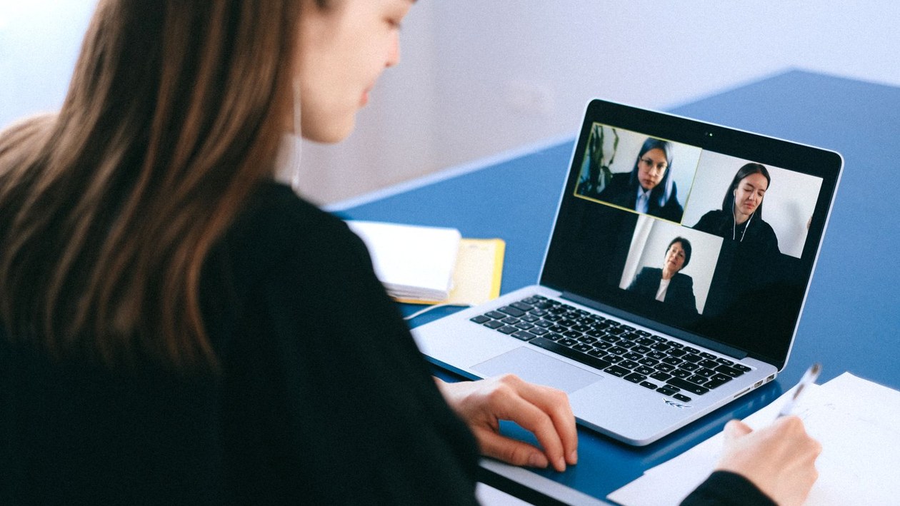 A woman seen from behind taking notes on paper while taking part in a video call with three people on her laptop screen.